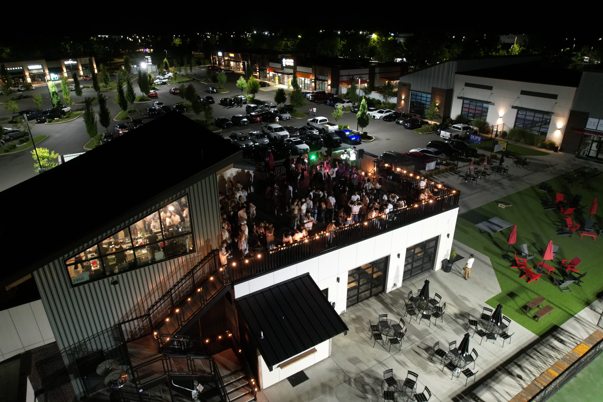 Aerial night view of a packed rooftop party at 23 Kitchens in Lacey, Washington, surrounded by a near-empty parking lot and quiet retail buildings.